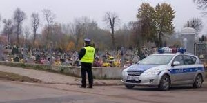 policjant obok radiowóz oznakowany, w oddali cmentarz