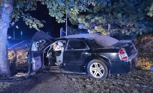 rozbity czarny samochód marki Chrysler po uderzeniu w drzewo, w oddali straż pożarna