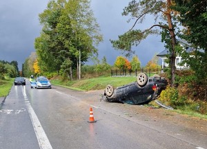 samochód marki BMW odwrócony na dachu po wjechaniu do rowu i uderzeniu w przepust, w oddali radiowóz i pojazd czekający na przejazd