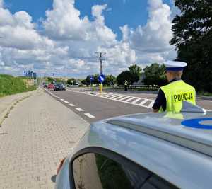 radiowóz, obok policjant odwrócony w stronę jezdni, którą przejeżdżają samochody