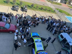 plac przed komendą policji  a na nim policjanci prowadzą pogadankę z młodzieżą, widać także wóz strażacki, i pojazdy policji i straży granicznej