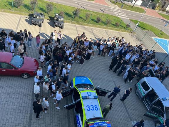plac przed komendą policji  a na nim policjanci prowadzą pogadankę z młodzieżą, widać także wóz strażacki, i pojazdy policji i straży granicznej