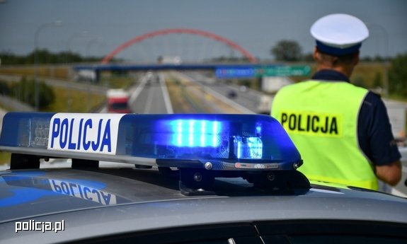 policjant ruchu drogowego obserwuje drogę, za nim sygnały świetlne radiowozu