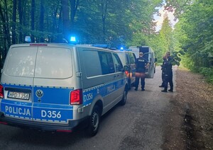 dwa radiowozy z włączonymi sygnałami świetlnymi, obok policjanci, w oddali zatrzymany biały bus