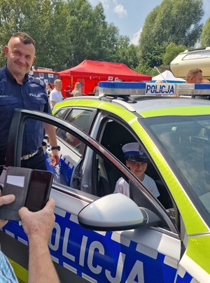 policjant obok radiowozu, na przednim siedzeniu chłopiec w czapce policjanta