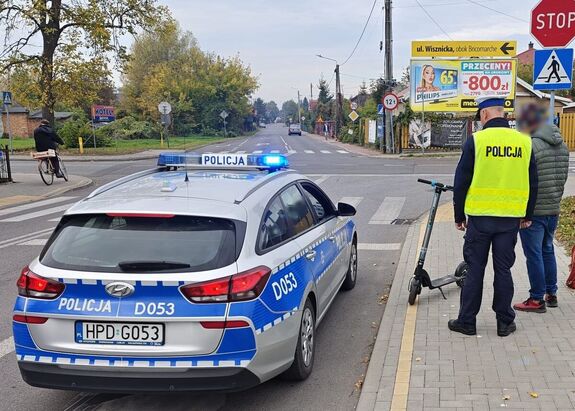 policjant kontroluje kierującego hulajnogą, obok radiowóz z włączonymi sygnałami świetlnymi